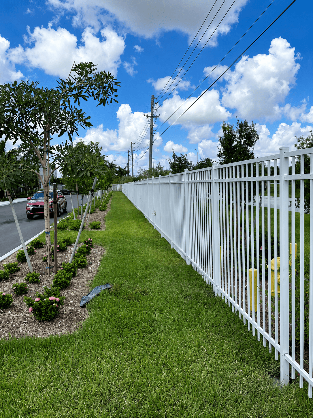 White flat-top ornamental metal fence along a landscaped roadside perimeter