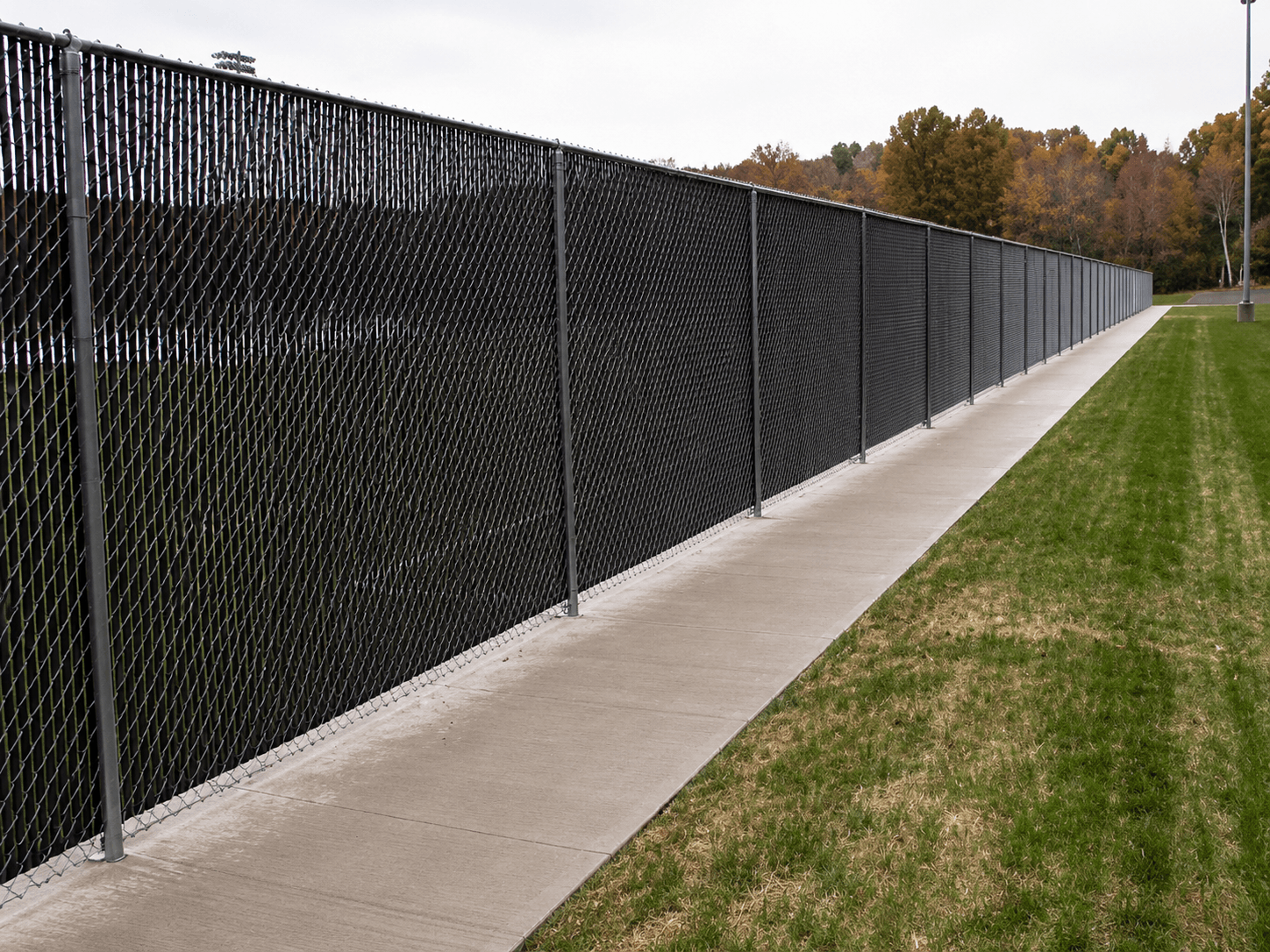Chain-link perimeter fence with black privacy slats along an athletic field walkway